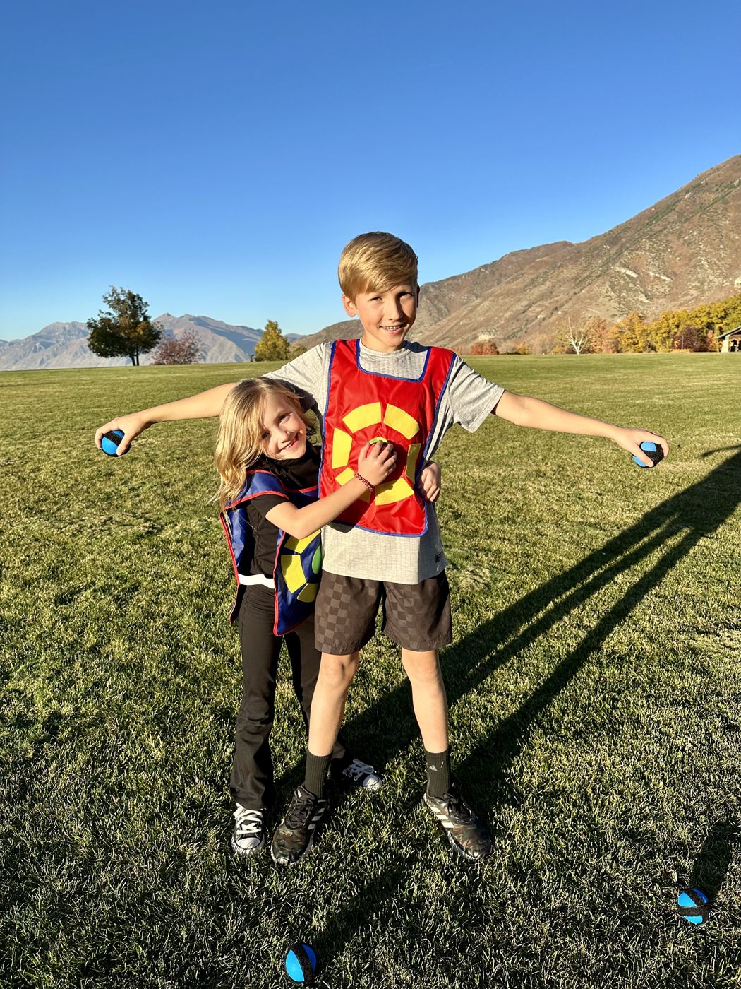 A boy holds out two sticky balls while his sister hugs him in a park below the mountains