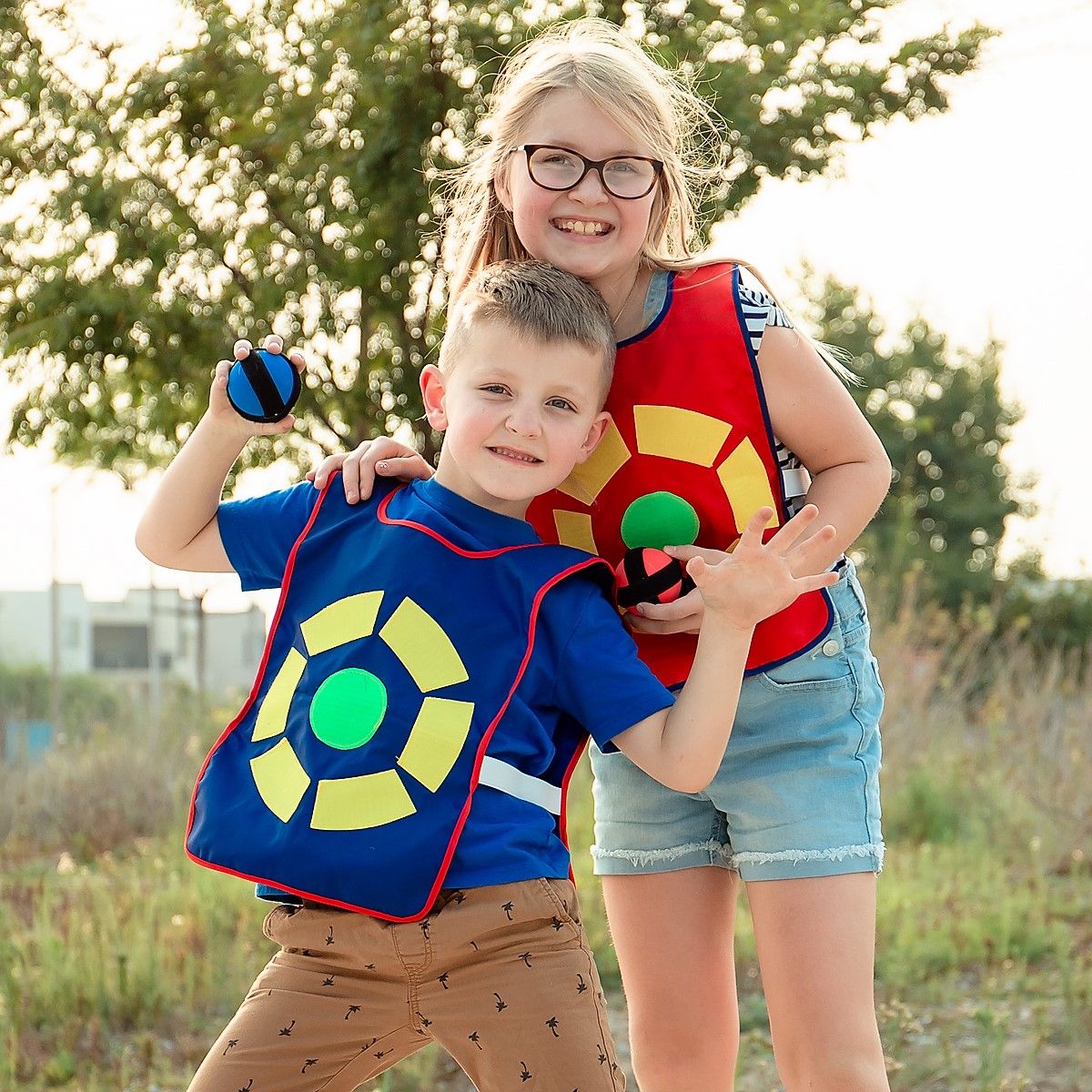 A smiling boy in a blue target vest winds up to throw a sticky ball while his sister in a red vest hugs him