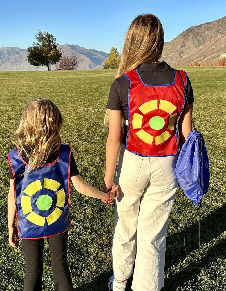 Two sisters in blue and red vests walk across a park holding hands, one carrying the blue drawstring bag